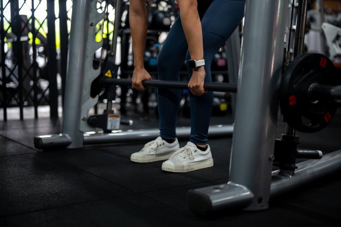 A woman with white shoes and blue leggings does Smith machine deadlifts.