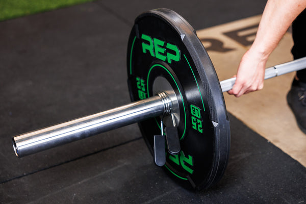 Close-up: Athlete grips a barbell loaded with 25 lb REP Sport Bumper Plates 2.0 and secured with a spring clip.
