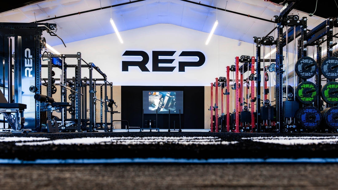 The second floor of REP's Colorado Showroom with a backlit REP logo on the wall, flanked by red and black power racks and stacked bumper plates.