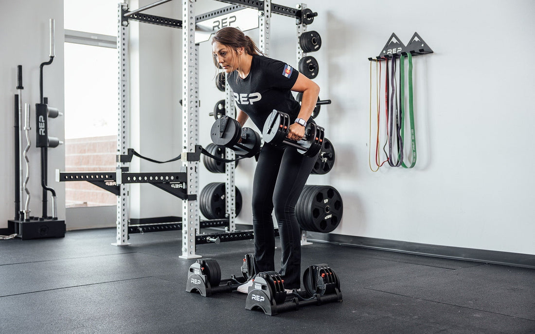Woman performing bent-over rows with the QuickDraw™ Adjustable Dumbbells. A REP power rack is behind her, with resistance bands on the wall.