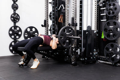 Person performing bench press with a barbell and weights off the front of the Summit™ All-In-One Ares™ 2.0 Wall-Mounted configuration in a gym setting.