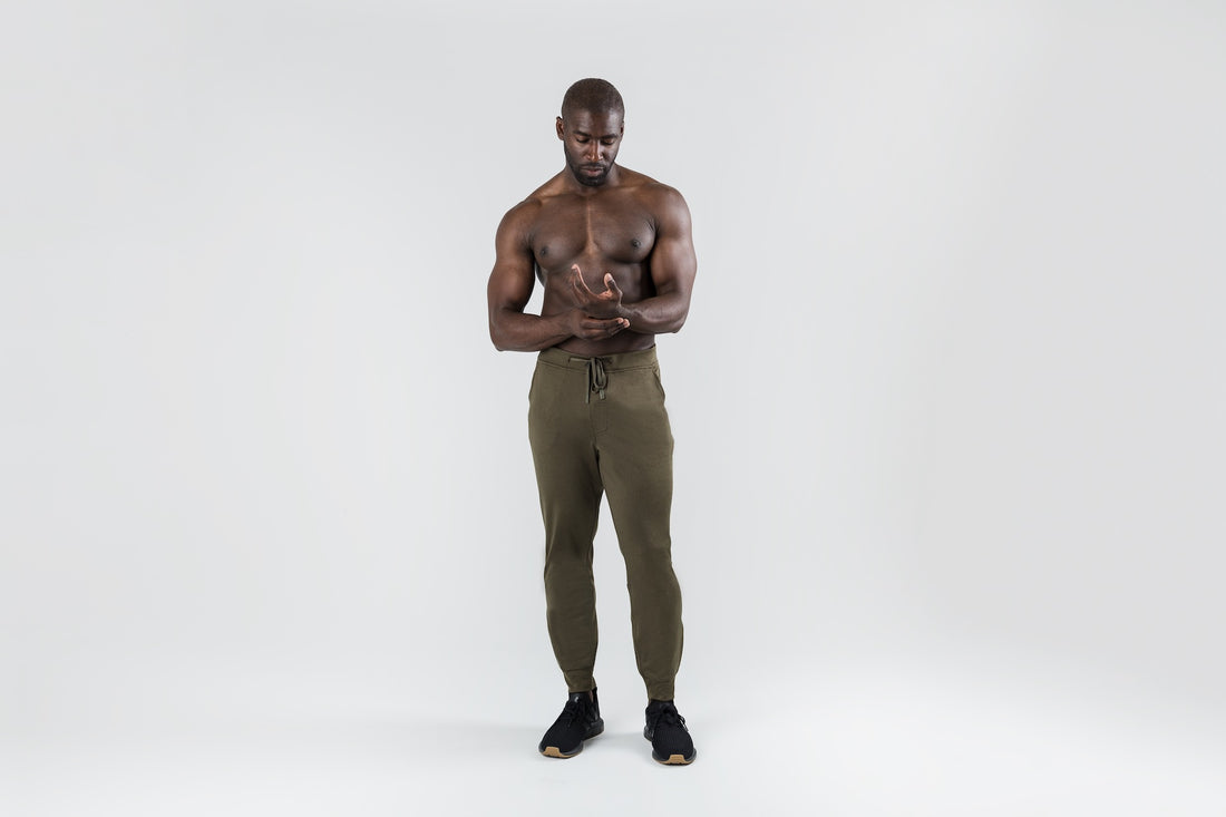 The REP Fitness Men's Attis Joggers in Heather Olive front, shirtless, adjusting drawstring