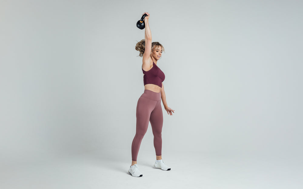 Side shot of an athlete holding a kettlebell over their head while wearing the REP Rhea Tank in Wine.