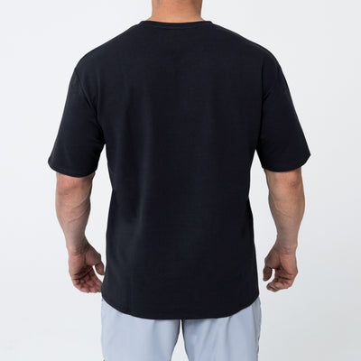 Men's Magnis Heavyweight T-Shirt in Black on person with grey shorts on a white background. Back profile. 