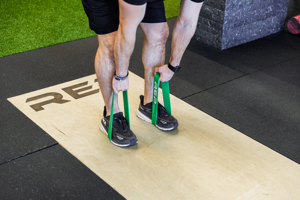 Lifter using green short resistance bands around his feet to perform banded deadlifts.