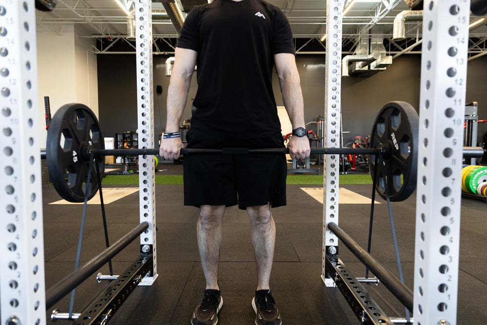 Lifter performing deadlifts inside the rack using a bar loaded with a pair of 45lb plates and also black short resistance bands attached via band pegs for added resistance.