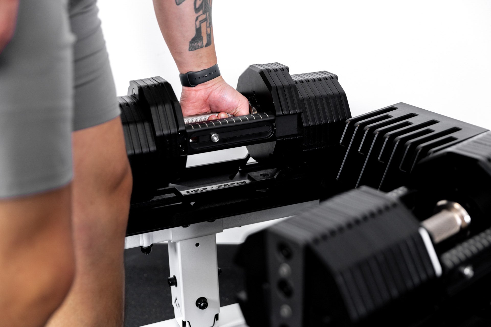 Athlete lifting up REP® x PÉPIN™ adjustable dumbbells from a white REP® Adjustable Dumbbell Stand