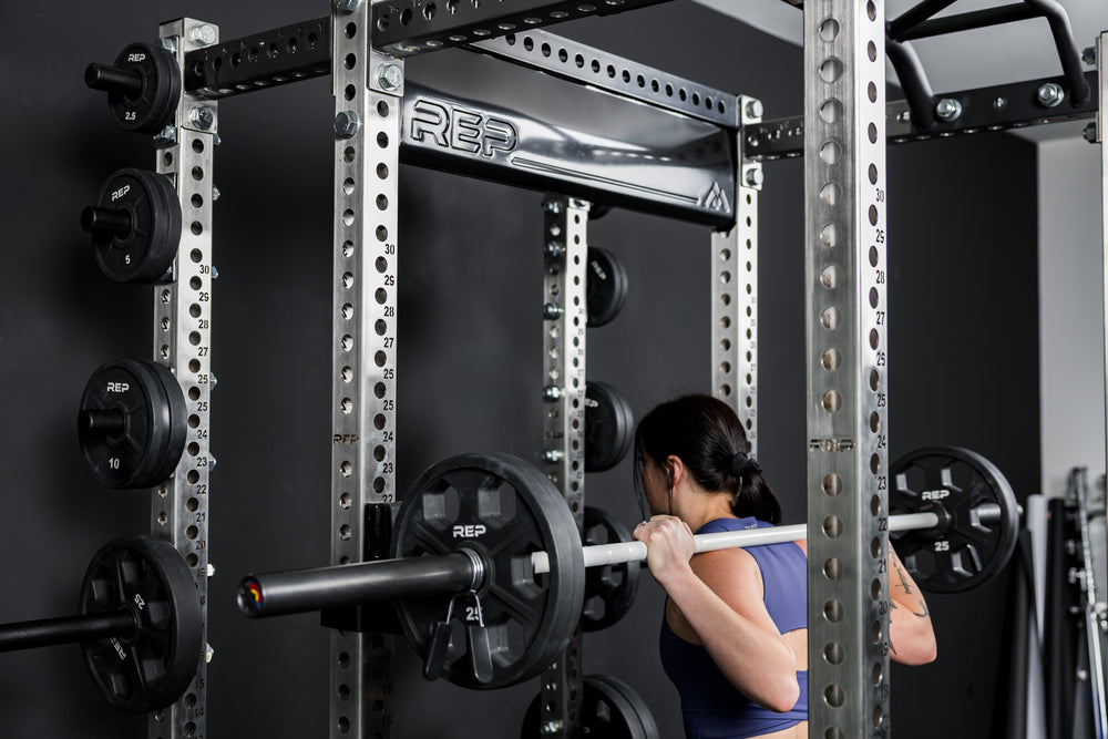 Person lifting weights with a power rack in a gym setting featuring the new stamped Logo Plate Crossmember 2.0