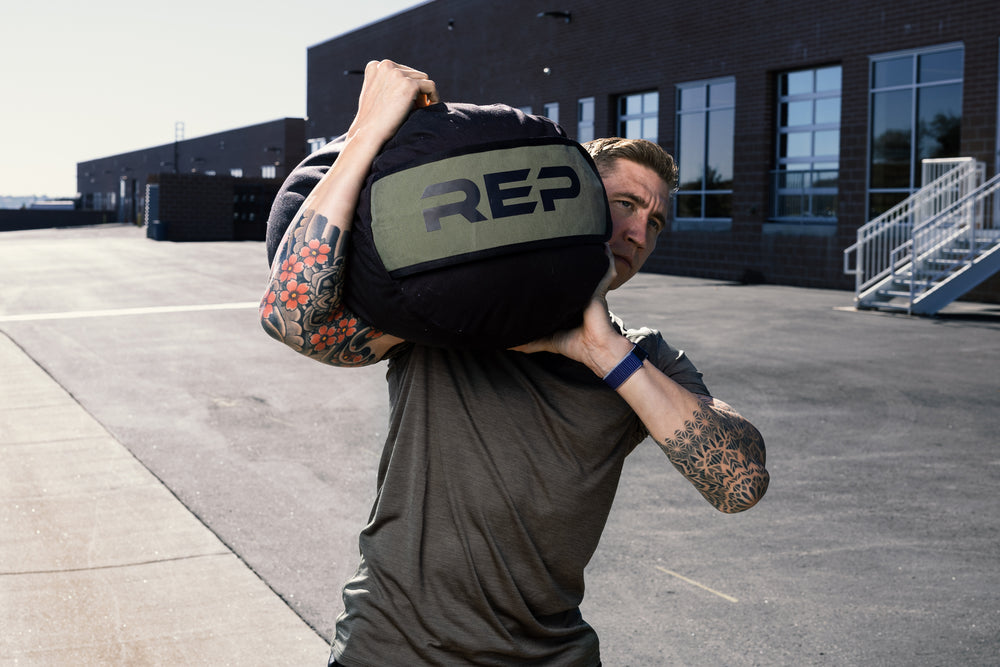 Person carrying a Rep Fitness  stone sandbag  on their shoulder outdoors.