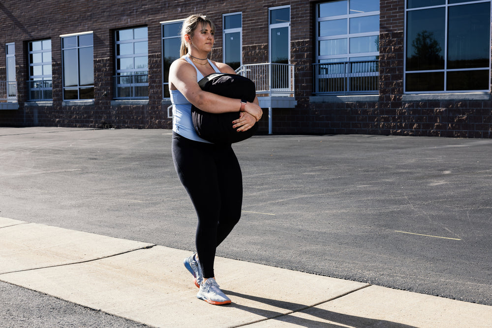 Woman performing a stone sandbag carry outdoors on a sidewalk with a building in the background
