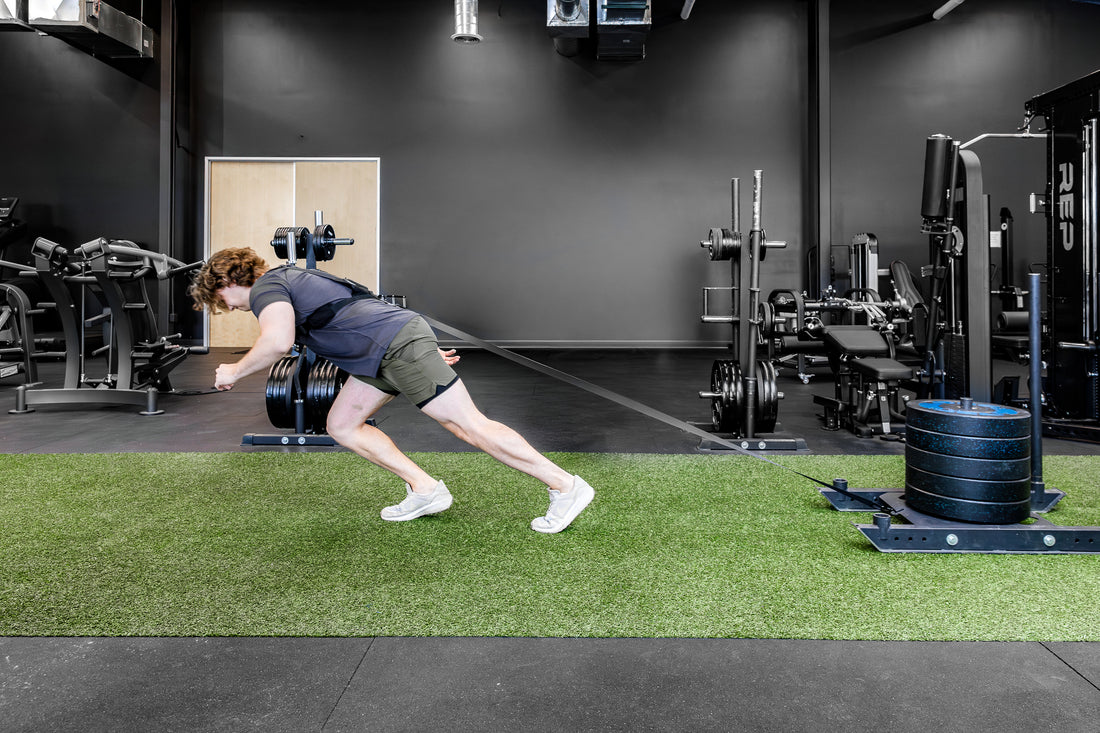 Person utilizing Sled Harness to pull a heavy sled on turf in a gym setting.