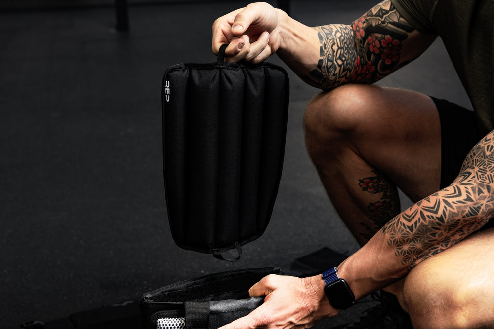 Person with tattoos holding a black soft weight vest plate by loop on the bottom in a gym setting