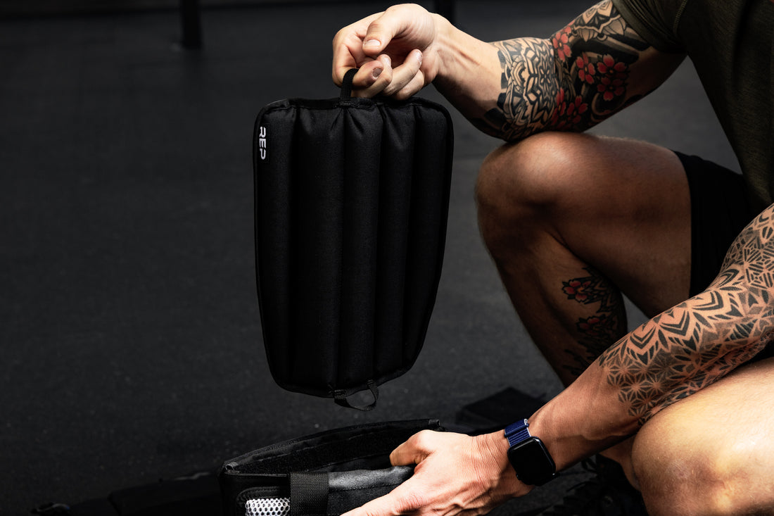 Person with tattoos holding a black soft weight vest plate by loop on the bottom in a gym setting