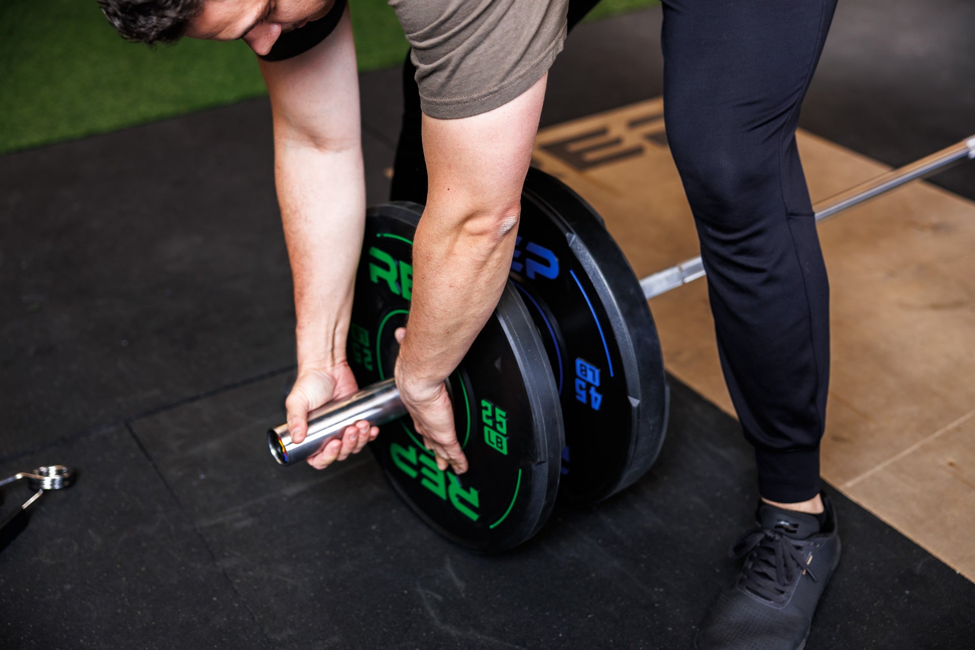 High-angle: Athlete adds a 25 lb REP Sport Bumper Plate 2.0 to a barbell already loaded with a 45 lb plate