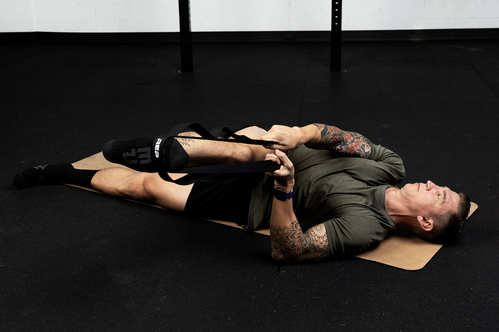 Person lying on a black mat with stretching bands around their foot and leg crossed over in a gym setting.