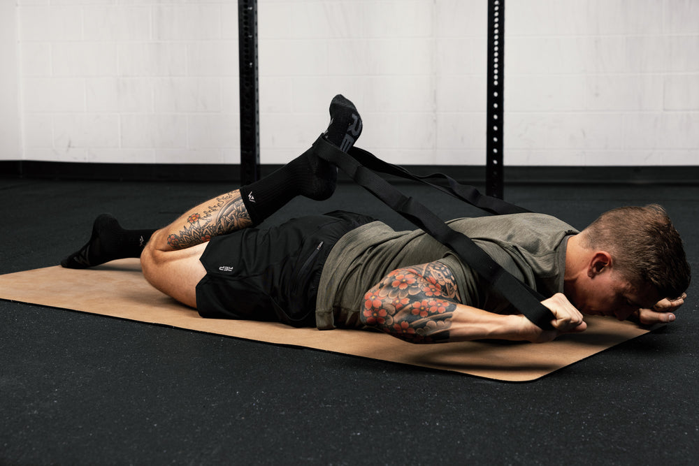 Person lying on a  yoga mat in a gym setting with visible tattoos using stretching band for quad