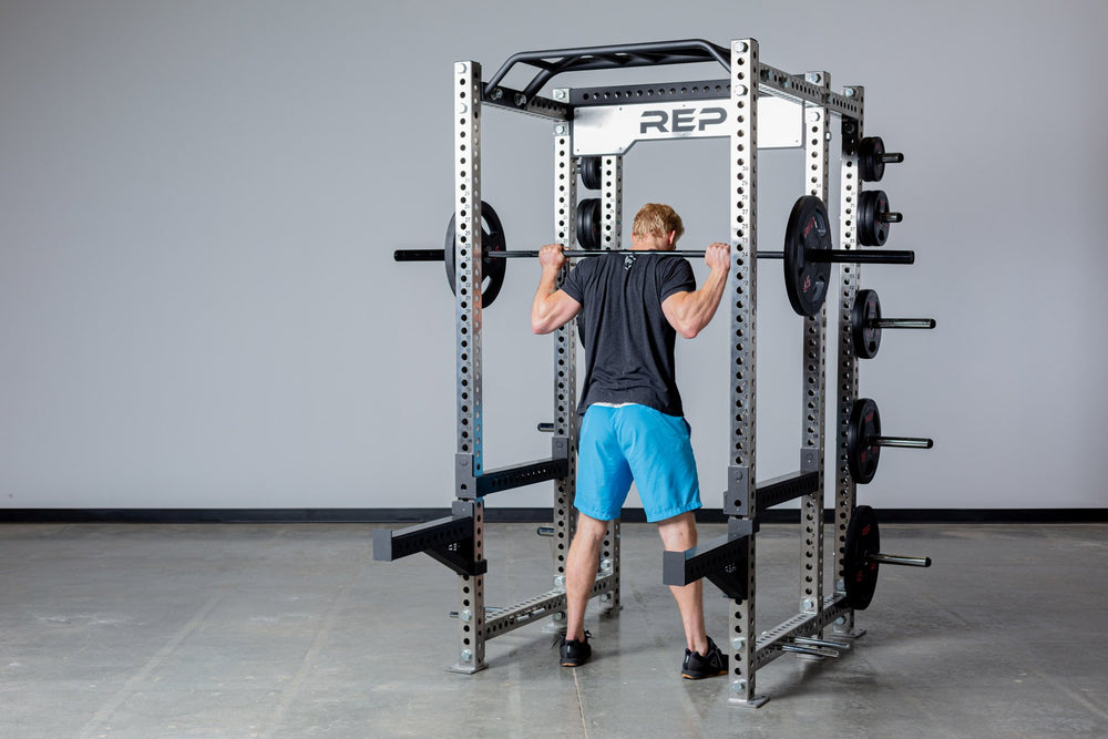 Lifter standing with a barbell loaded with a pair of 45lb Rubber Coated Olympic Plates in the back rack position inside of a 6-post PR-5000 power rack.