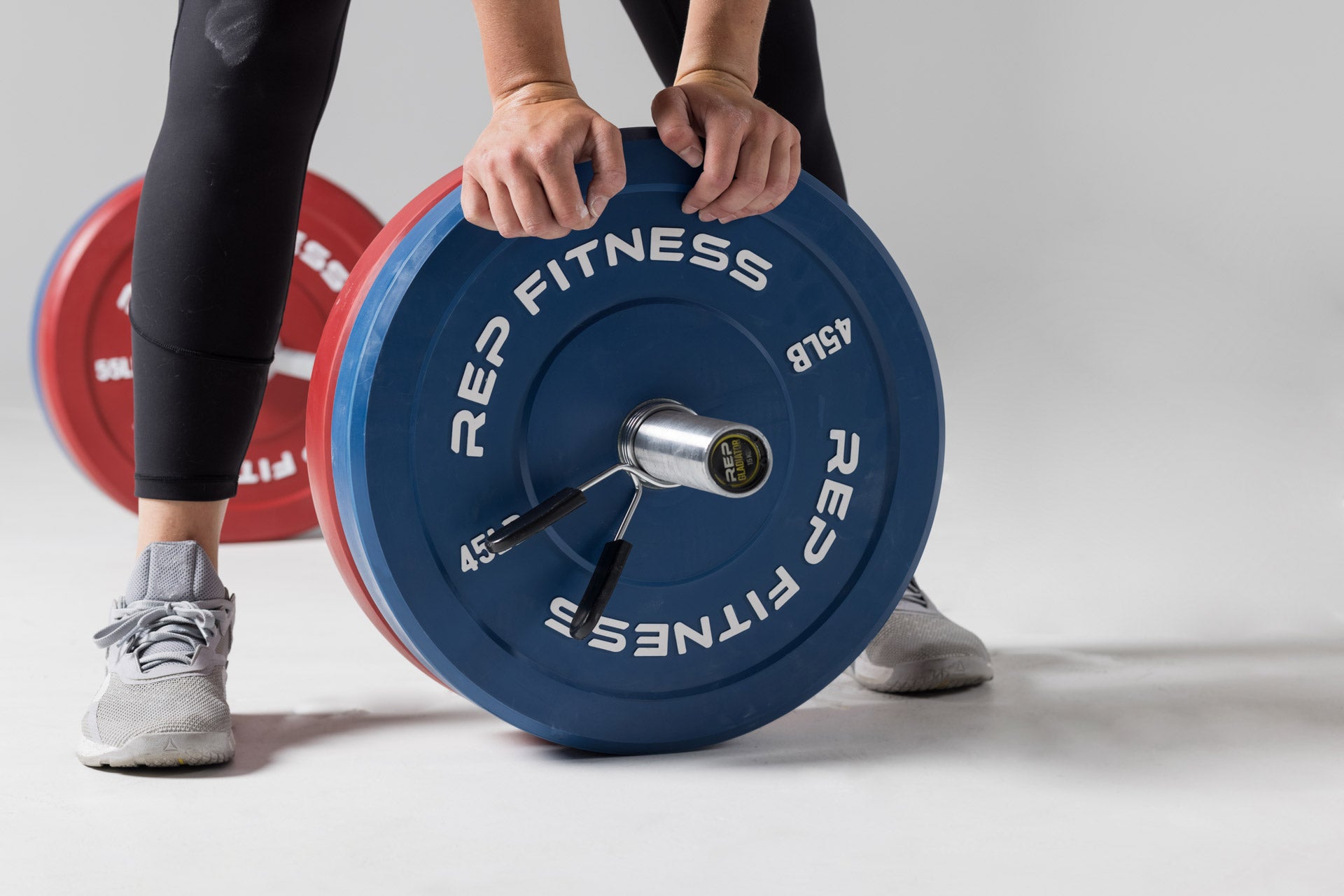 Close-up view of a lifter standing over a barbell on the ground after having just loaded it with a pair of red 55lb and blue 45lb colored bumper plates.
