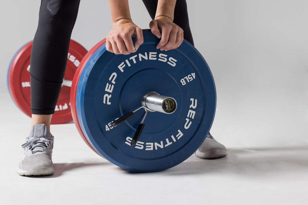 Close-up view of a lifter standing over a barbell on the ground after having just loaded it with a pair of red 55lb and blue 45lb colored bumper plates.