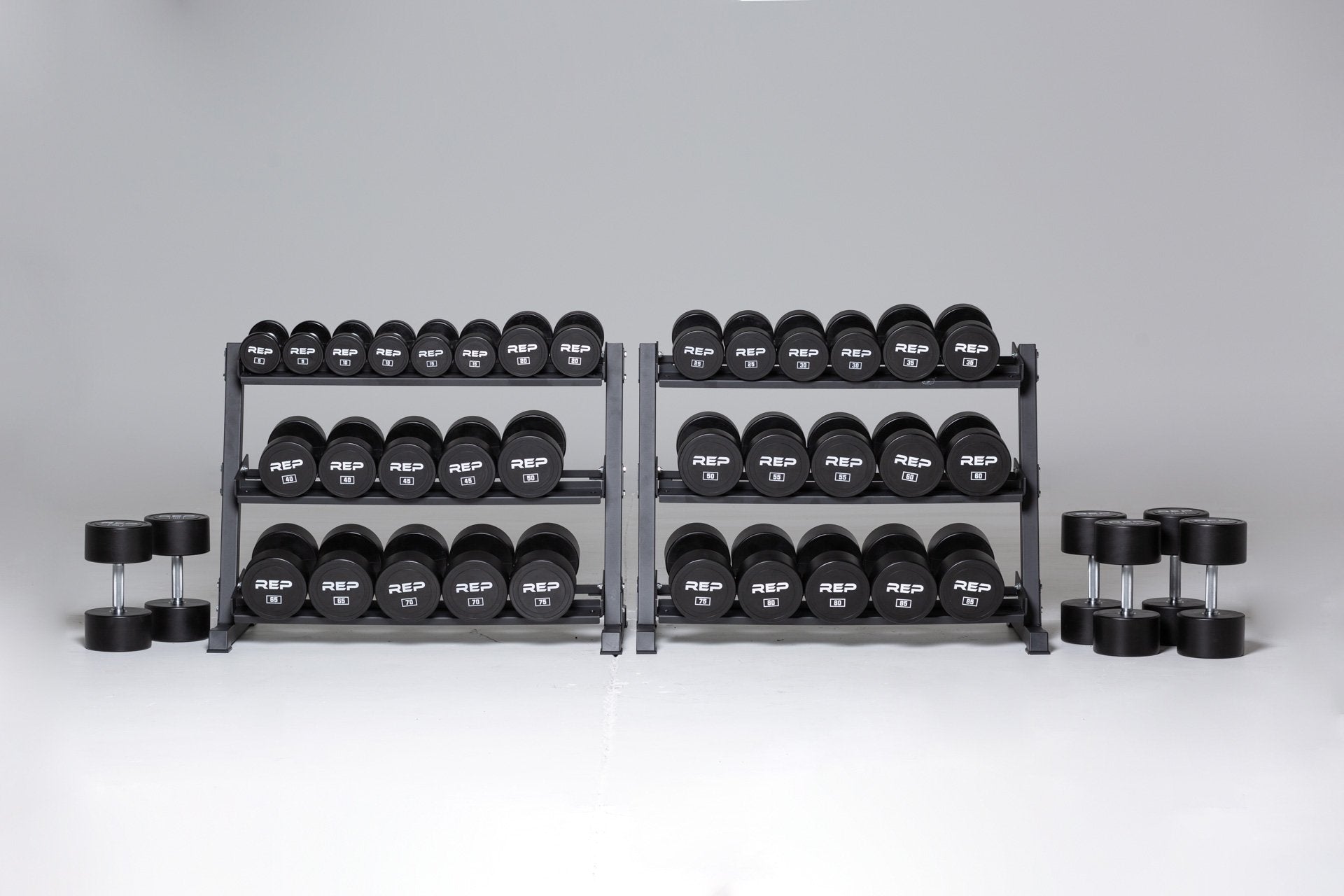 5-100lb Urethane Dumbbell Set stored on two REP Dumbbell Racks with the 90, 95, and 100lb pairs being stored on the ground next to the racks.
