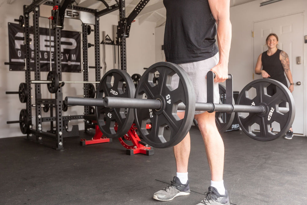 Lifter in a garage gym standing while holding a pair of REP Farmer's Walk Handles, each loaded with a pair of 45lb Urethane Coated Equalizer Plates.