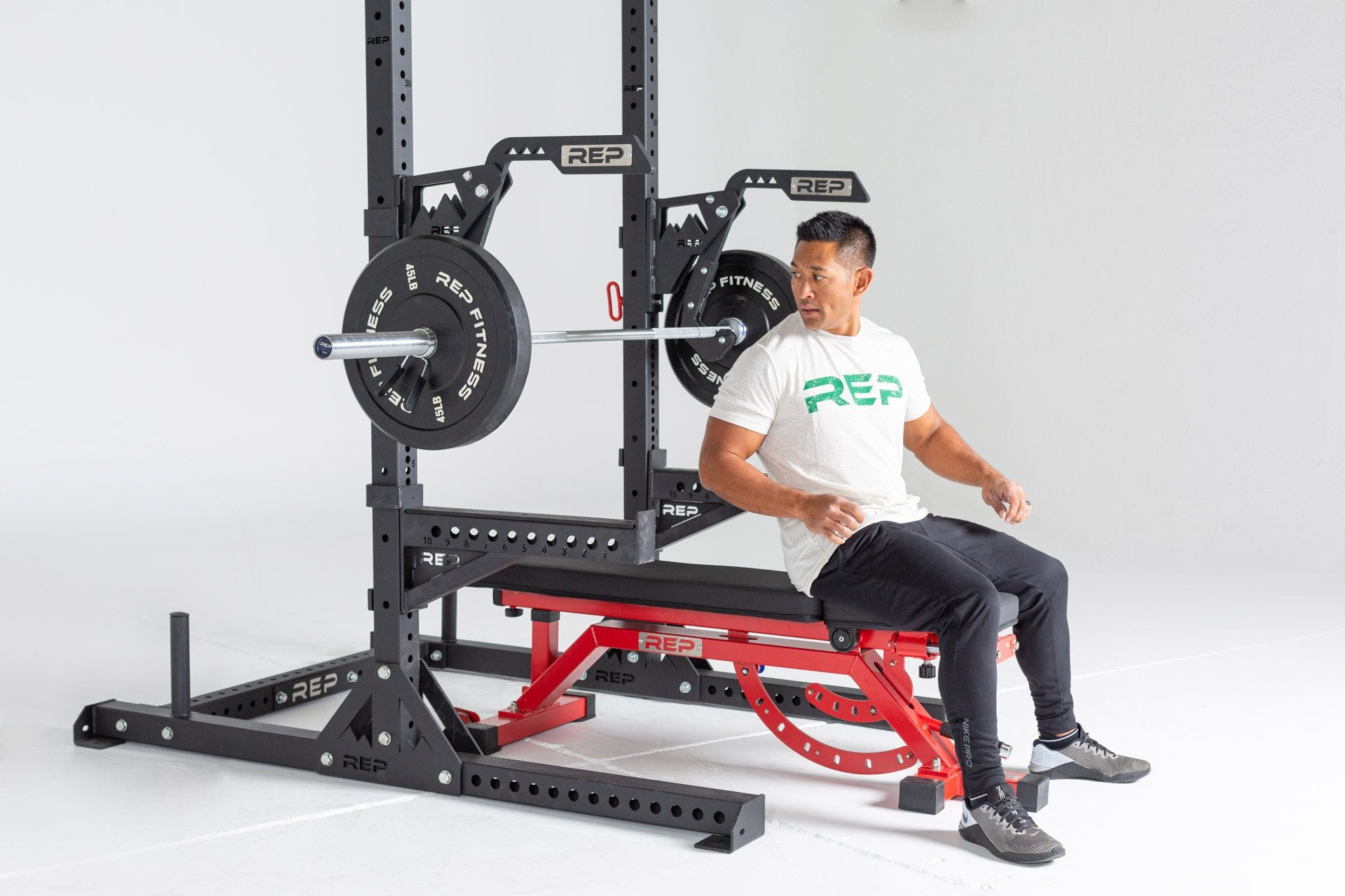 Lifter sitting on an AB-5000 Bench inside a REP Squat Rack with REP Monolift Arms and Spotter Arms getting ready to lie back and perform a bench press with a barbell loaded with a pair of 45lb Black Bumper Plates.