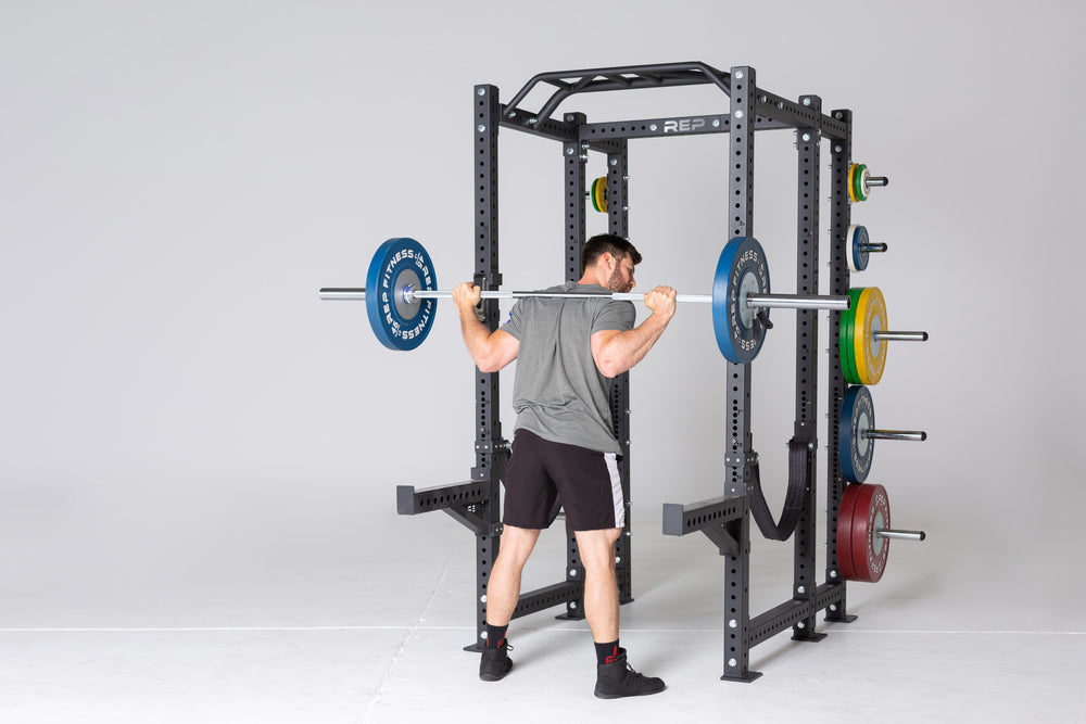 Lifter with a barbell loaded with 45lb Competition Bumper Plates in the back rack position preparing to squat outside of a PR-4000 rack with spotter arms, strap safeties, and weight horns storing Competition Bumper Plates (LB) and change plates.